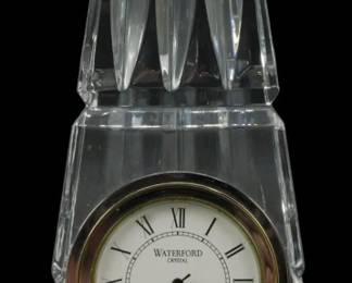 908 - Waterford Crystal Obelisk Desk Clock (Description): Waterford Crystal obelisk desk clock  featuring a distinctive architectural tower  design. The cut crystal body showcases  vertical faceted panels rising to a pointed  pyramidal top. The clock face displays the Waterford logo, Roman numeral markers, and a  gold-tone bezel set into the front of the  obelisk base. Used as is electric/electronic/mechanical  item. No guarantee all components, untested.