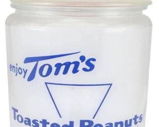 1005 - Tom's Peanuts Store Jar 10.5"x6.5" (Description): Tom's Peanuts store jar measuring 10 1/2" x  6.5". This clear glass counter jar features  blue printed advertising reading "enjoy Tom's  Toasted Peanuts Delicious" with the  signature Tom's triangle logo. The jar retains its original white lid with Tom's  finial. Text along the bottom reads "Prop.  TOM'S Distbr. — Not to be sold." A classic  piece of mid-century American advertising and  general store memorabilia.