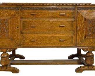 7779 - Vintage English Carved Oak Sideboard Buffet (Description): Antique carved oak sideboard buffet featuring  beautiful quarter-sawn oak construction  throughout. The piece showcases carved  grapevine motifs on the two side cabinet  doors, with three center drawers fitted with wooden pulls. The top edge displays an ornate  carved border detail. The buffet rests on an  impressive trestle-style base with turned  and fluted bulbous supports, decorative  scroll brackets, and a lower stretcher