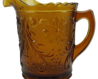 1071 - Vtg Indiana Glass Amber Sandwich Pitcher 7.5" (Description): Vintage Indiana Glass amber Sandwich pattern  pitcher measuring 7.5 inches tall. This  pressed glass pitcher features the classic  Sandwich pattern with embossed floral and  scroll designs against a stippled background. The pitcher has a scalloped rim with a  shaped pour spout and an applied handle. The  rich amber coloring is consistent throughout.