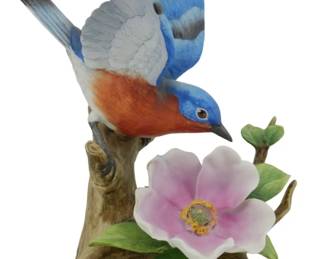 1073 - Andrea by Sadek Bluebird Porcelain Figure 7" (Description): Andrea by Sadek porcelain figurine depicting  a bluebird perched on a naturalistic tree  branch with wings raised. The bird features  hand-painted plumage in vibrant blue,  rust-orange breast, and gray-white accents. A delicate pink wild rose blossom with yellow  center and green leaves adorns the base.  Measures 7 inches.