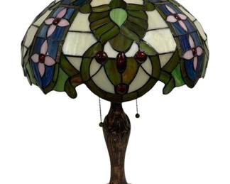 1511 - Tiffany Style Stained Glass Table Lamp 24" (Description): Tiffany style stained glass table lamp  featuring a dome shade with a floral and leaf  motif. The leaded glass shade incorporates  green leaves, blue accents, pink flower  petals, and deep red cabochon jewels against a cream background, with a scalloped edge.  The lamp sits on a decorative cast metal base  with an antiqued bronze finish and Art  Nouveau-inspired design. Features dual pull  chain operation. Measures 24 inches tall.