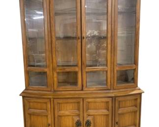 7585 - Mid-Century American of Martinsville Cabinet (Description): Featuring a lighted upper display section  with glass panel doors and sides, ideal for  showcasing fine china and collectibles. The  upper portion has four glass-front doors with  beveled frames. The lower buffet base features two cabinet doors with raised panel  detailing and decorative brass drop pulls,  providing enclosed storage and lined  silverware drawer. The piece has canted  corners and a warm medium oak finish.