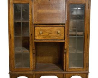 7762 - English Oak Secretary Cabinet w/ Leaded Glass (Description): Antique oak secretary cabinet featuring a  drop-front writing desk, single drawer with  brass knobs, and flanking display cabinets  with leaded glass doors. The piece showcases  quarter-sawn oak construction with attractive grain patterns throughout. The central  section includes an open storage compartment  below the drawer. Resting on a decorative  scalloped base with bracket feet. Missing 1  pane of glass. Measures 45" x 40" x 12".