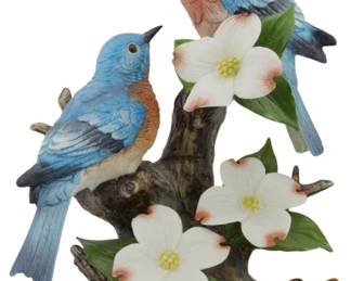 1087 - Andrea by Sadek Porcelain Bluebirds Figure (Description): Andrea by Sadek porcelain figurine featuring  a pair of Eastern Bluebirds perched on a  naturalistic tree branch adorned with white  dogwood blossoms and green foliage. The birds  are finely detailed with vibrant blue plumage on their wings and backs,  russet-colored breasts, and delicate facial  features. The sculptural base depicts a  gnarled brown branch rising from an earthtone  ground, with realistic dogwood flowers