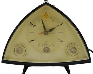 789 - MCM Silvertone Magic Glow Wedge Clock Radio (Description): Mid Century Silvertone transistor clock radio  featuring a distinctive triangular design  with black casing and tripod legs. The solid  state unit includes a cream-colored dial face  with stylized numerals, analog clock hands with red second hand, and integrated controls  for volume (soft/loud), alarm settings, and  AM radio tuning. Features wake to music and  alarm functions. Measures 8 inches by 8  inches by 4 inches.