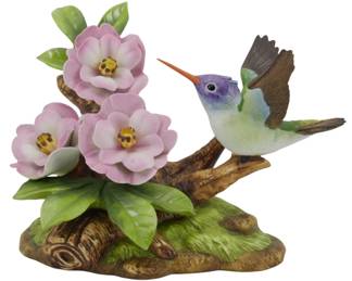 1083 - Andrea by Sadek Porcelain Hummingbird Figure (Description): Andrea by Sadek porcelain figurine depicting  a Violet Crowned Hummingbird perched on a  naturalistic branch with pink flowers and  green foliage. The hummingbird features  delicate hand-painted details including a violet crown, green and white body, brown  wings, and distinctive orange beak. The  sculptural base incorporates textured bark  and moss elements. Measures 5.5 inches.