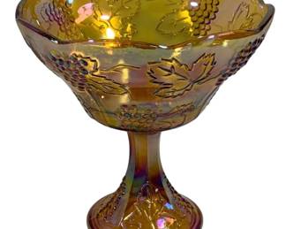 1550 - Amber Carnival Glass Grape Pattern Compote (Description): Carnival glass compote in amber iridescent  finish featuring a grape and leaf pattern.  The bowl has a scalloped edge and sits atop a  pedestal base with matching decorative  motifs and beaded details. Measures 8.5" x 8.5".