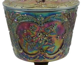 675 - Fenton Carnival Glass Strawberry Compote (Description): Vintage Fenton Wild Strawberry carnival glass  covered compote. This striking pedestal jar  features the iconic Wild Strawberry pattern  with raised strawberries, blossoms, and  foliage in decorative cartouches. The iridescent finish displays beautiful rainbow  hues of purple, green, gold, and pink typical  of quality carnival glass. The piece  includes a fitted lid with a ribbed finial  handle. Measures 9 1/2" x 6".