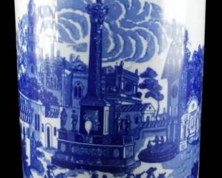 1038 - Blue & White Cylindrical Umbrella Vase 17" (Description): Large blue and white cylindrical planter  featuring a transferware-style scenic design  depicting a classical European town square  with a central monument or column, figures on  horseback, pedestrians, and architectural buildings. The rim is decorated with a  complementary border pattern featuring  landscape vignettes and floral motifs.  Measures 17 inches in height.