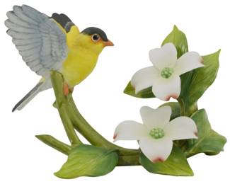 1090 - Andrea by Sadek Porcelain Goldfinch Figure (Description): Andrea by Sadek porcelain figurine featuring  an American Goldfinch perched on a branch  with dogwood blossoms. The bird is depicted  with wings spread, showcasing detailed yellow  plumage, black cap, and gray-blue wings. The naturalistic base includesgreen leaves and  two delicate white dogwood flowers with  pink-tipped petals. Measures 5 inches.