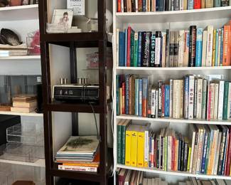 A 1970s vintage Sony Digimatic flip clock radio, a collection of black zippered cases and tech organizers, a dark brown vertical four-shelf modular shelving unit, a five-shelf bookcase, lots of collectible books on art, interior design, history, remodeling, military history, architecture, automobiles, travel;