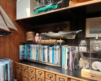A vintage fire boat ship floodlight with a red metal housing and ribbed Top Spredlite glass lens, a vintage oak 18-drawer card catalog or apothecary unit cabinet with brass label frames and pulls; great collection of books about sailing, ships, seafaring warfare, nautical history; model ships such as the Revell Cutty Sark, a wooden model of a ship, likely a clipper, with intricate rigging and a solid wood hull, and a wooden sailboat model (schooner or yacht design)