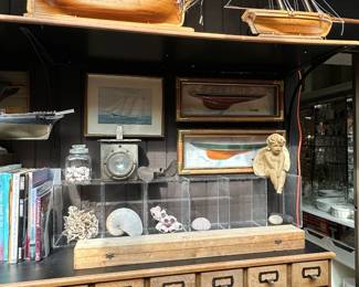 A vintage oak 18-drawer card catalog or apothecary unit cabinet with brass label frames and pulls; a wooden model of a ship, likely a clipper, with intricate rigging and a solid wood hull; a wooden sailboat model (schooner or yacht design), acrylic storage or display unit with seashells and coral; framed nautical prints, a vintage WWII-era U.S. Navy battle lantern, a vintage Kiraku & Co. "Fresh Water Set" split bamboo fly fishing rod in wooden case, and a cast resin cherub shelf sitting figurine