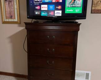 Flat-screen smart TV displayed on a tall, dark wood five-drawer chest. The TV is powered on and appears to be in working condition. The chest provides generous storage and features a rich wood finish with metal drawer handles.  The TV and chest are being sold separately unless otherwise noted.