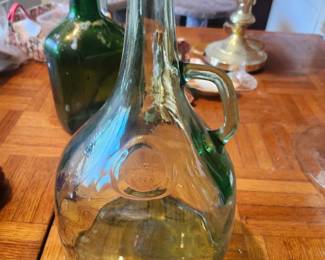 Description  Vintage green glass wine or liquor bottle featuring a molded side handle, embossed circular medallion marked "3/4 L 1950", and a tall narrow neck. This European-style utility bottle makes an attractive decorative piece for farmhouse, rustic, or wine cellar décor.  Its classic shape and rich green glass make it perfect for display, dried flowers, or as part of a vintage bottle collection.  Features:  Embossed "3/4 L 1950" medallion Original molded glass handle Rich green glass Decorative European-style bottle Great for farmhouse, kitchen, bar, or wine cellar décor Condition Good vintage condition No visible cracks or major chips Light wear consistent with age Displays very well Please review photos for complete condition Estimated Value Estate Sale Price: $20 Online Marketplace Value: $25–45 Antique Shop Retail: $35–60  Buyer pays shipping or local pickup available