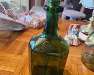 Vintage emerald green glass bottle featuring an embossed circular medallion on the front and a distinctive square-body design with rounded shoulders. The bottle is unmarked otherwise and would make an excellent addition to a bottle collection, primitive farmhouse décor, bar display, or floral arrangement. Clean condition with no major chips or cracks visible from the photos. Displays beautifully.  Approximate age: Mid-20th century (exact manufacturer unknown).  Condition:  Very good vintage condition No visible cracks or major chips Light age-related wear consistent with vintage use Sold as pictured  Estimated Value:  Estate Sale Price: $15 Online Marketplace Value: $20–35 Antique Shop Retail: $30–45  Pickup/Shipping: Buyer pays shipping or local pickup available.