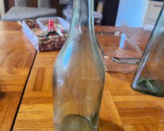 Vintage Embossed Aqua Glass Soda Bottle  Authentic vintage embossed aqua glass soda bottle featuring classic early 20th-century styling and raised lettering. This collectible bottle displays the beautiful light aqua glass color sought after by bottle collectors and makes an attractive display piece for farmhouse décor, antique collections, or man caves.  Perfect for collectors of antique bottles, advertising memorabilia, or vintage Americana.  Features:  Original embossed glass bottle Light aqua glass Raised lettering and molded details No cap Early machine-made bottle Approximate Age: 1900–1925  Condition: Good antique condition with age-related wear, light interior staining, and minor surface scratches consistent with age. No major cracks or chips visible. Please review all photos for condition.  Estate Sale Price  $20.00  Shipping  Buyer pays actual shipping. Local pickup is also available.