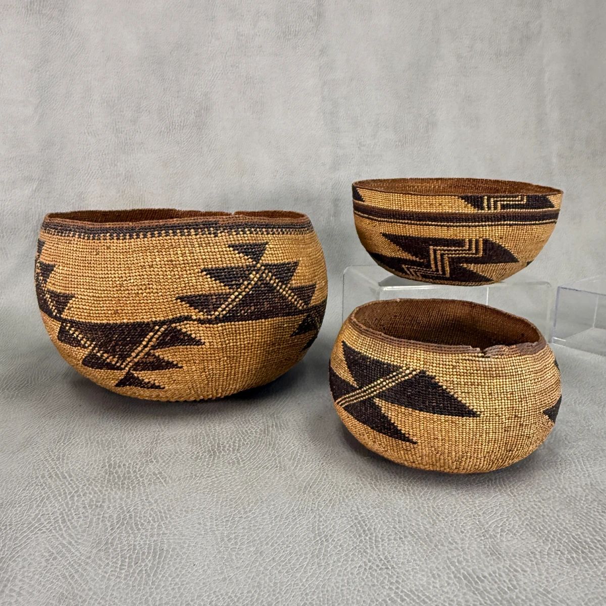 A northern California Native American (possibly Hupa, Yurok or Karuk) large twined basket with geometric designs of stepped triangular motifs and linear bands, approx 9-3/4”W x 6”H; an Indigenous peoples of Northern California traditional twine-woven basket cap or hat with stacked geometric zigzag elements on the side and repeating at the rim above a black and brown band, approx 7”W x 4”H; and a Shagton Group Pit River California woven natural fibers basket with pattern frequently interpreted as representation of geese in flight or stacked flint blades; approx 7” wide
