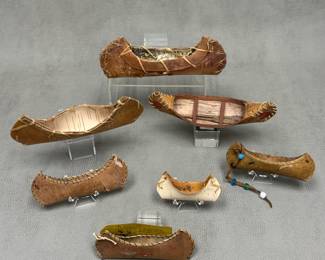 A collection of seven handcrafted miniature birch bark canoes, featuring traditional stitched bark hulls and wooden framing details; largest is 9 inches long, smallest is approx. 3 inches long.