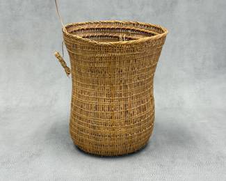 A handwoven wicker storage basket, cylindrical with a slightly flared rim and rounded base, with an open-work or spaced-weft weave style.  6-1/2"W x 9"H.