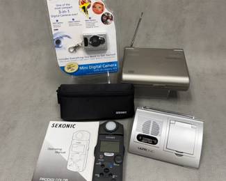 A black Sekonic Prodigi Color C-500 or C-500R Color Meter with a digital display, control buttons, and a rotating dial, with original manual and carrying case; an Innovage Mini Digital Camera 3-in-1 Keychain Accessory Kit in original package; an Ultronic Desktop AM/FM Radio with Calendar and Clock (Model T524)
