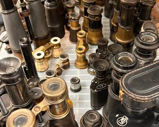 A large selection of antique and vintage optical instruments, mostly consisting of 19th- and early 20th-century brass field glasses, marine binoculars (jumelles marine), opera glasses, and pocket telescopes. Many are made from polished brass, blackened steel, and wrapped in materials like genuine leather or mother-of-pearl.