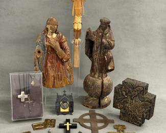 A collection of religious sculptures, jewelry, charms, and crucifixes including a vintage Mexican folk art handcrafted woven straw crucifix with detailed straw figure of Jesus Christ on a simple wooden cross; an antique European or Latin American polychrome carved wooden devotional sculpture depicting a robed saint or the Virgin Mary; an antique carved wooden santo (devotional figure), likely depicting a friar or saint; crucifix pendant necklaces; and a vintage Mexican folk art milagros cross sculpture created by artist Estela Ogazón (1966–2009)