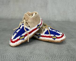 A child’s sized pair of traditional Native American beaded leather moccasins, likely Plains Indian or Lakota Sioux-style; with glass seed beadwork featuring a central vertical white stripe flanked by bold blue fields and red-and-white curved border motifs on soft suede hide. Integrated leather tie laces through the cuff fold.