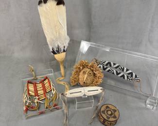 Native American items including a handmade smudging fan crafted using feathers attached to a leather-wrapped handle; a pair of traditional Inuit snow goggles (iggaak or ilgaak) carved from bone or antler with leather or sinew side straps; a Native American bone and bead breastplate; and a coiled woven Native American basket, with cylindrical, flat-topped lidded shape and intricate polychrome geometric or floral star motif woven into the structure.