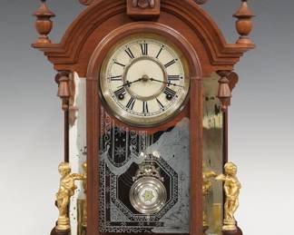 A late 19th century Waterbury Oxford model mirror side shelf clock.  8-day time and strike movement with papered metal dial and Roman numerals.  Walnut case with turned finials features a carved arched crest with Liberty mask above an arched door with stenciled glass flanked by mirrored pilasters and cast cherub figures on a rounded base.  Refinished with minor wear, dial wear, replaced cherubs, running when cataloged.  23" high.  ESTIMATE $100-150