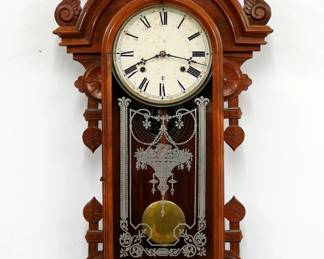 A late 19th century Gilbert Columbia model wall clock.  8-day time and strike movement with painted metal dial and Roman numerals.  Eastlake Period Walnut case with carved detail and applied Colombia mask, features a shaped crest with turned finials over an arched door with stenciled glass flanked by shaped ears over a drop with shaped apron and turned finials.  Paper label on back 10% intact.  Older refinishing with minor wear, re-papered dial with staining, running when cataloged.  35 3/4" high overall. ESTIMATE $400-600  