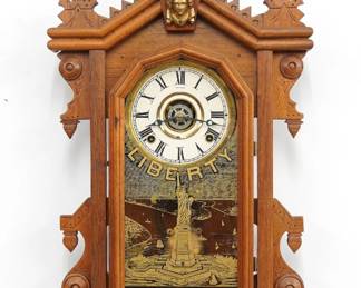A turn of the century Ingraham Liberty model Kitchen wall clock , retailed by Charles F. Adams, Erie, PA.  8-day time, strike and alarm movement with papered metal dial and Roman numerals.  Walnut case with turned finial and shaped crest with Liberty mask over and molded door with "Liberty" stenciled glass and a shaped drop.  Older refinishing with minor wear, running when cataloged.  25 1/4" high.  ESTIMATE $100-150