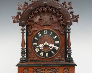 An early 20th century German Black Forest mantel Cuckoo table clock.  Good quality Brass 8-day time and strike movement with Cuckoo and Gong strike, wooden dial with applied Roman numerals.  Walnut case with Ebonized detail features an arched Grape leaf crest, turned columns and a molded base with carved detail.  Old finish with wear and touch-ups, small glued crest repairs, running when cataloged.  21" high.  ESTIMATE $400-600