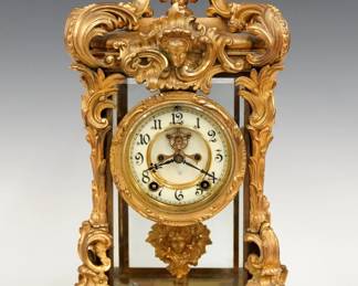 A turn of the century Ansonia Regal model crystal regulator. 8-day spring driven time and strike movement with two part porcelain dial, visible escapement and Arabic numerals with a cast mask pendulum. Louis XV Style Gilded Brass case with scrolled foliate detail, beveled glass doors and sides on a scrolled base with modified paw feet. Some wear, running when cataloged. 15 1/2" high overall. ESTIMATE $1,000-2,000