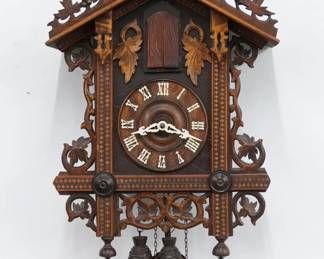 A turn of the century Black Forest Cuckoo wall clock.  8-day time and strike, three horn Trumpeter two weight movement with Brass cut-out plates and wooden dial with applied Roman numerals and filigree hands.  Mixed wood Railroad type case with hand carved detail features a foliate crest on a body with inlaid posts and grape leaf detail with turned finials.  Older finish with  minor wear, running when cataloged.  27" high plus weight drop.  ESTIMATE $800-1,200