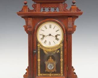A 19th century E. N. Welch Judic model Victorian shelf clock. 8-day time and strike movement with painted metal dial and Roman numerals, fancy pendulum with glass insert. Walnut case with shaped gallery top and turned finials above a single arched door with stenciled glass, flanked by fluted pilasters on a molded base. Paper label on back 60%. Older finish with minor wear, re-painted dial, running when cataloged. 19 3/4" high. ESTIMATE $300-400