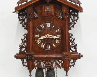 An early 20th century German Black Forest Cuckoo wall clock by Gordian Hettich & Son, Furtwangen.  30-hr Brass time and strike two weight movement with wooden dial and applied Roman numerals.  Walnut case with hand carved detail features a foliate crest on a body with molded posts and grape leaf detail with turned finials.  Paper label on back 80% intact.  Refinished with minor wear, running when cataloged.  26 1/2" high plus weight drop.  ESTIMATE $600-800