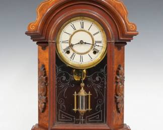 A turn of the century Waterbury kitchen shelf clock.  8-day time and strike movement with papered metal dial and Roman numerals.  Walnut case with gilded detail features a shaped crest and Liberty mask over an arched molded door with stenciled glass flanked by chamfered Burl sides with carved medallions and a shaped molded base.  Refinished with minor wear, minor dial wear, running when cataloged.  22 1/2" high.  ESTIMATE $100-150