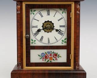 A late 19th century New Haven Cottage shelf clock. 30-hr spring driven time, strike and alarm movement with papered metal dial and Roman numerals.  Rosewood case with a single door, clear dial glass and reverse painted lower flanked by turned quarter columns on a simple molded base.  Interior paper label 80% intact.  Refinished with slight wear, re-papered dial, running when cataloged.  13" high.  ESTIMATE $100-150