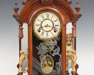 A late 19th century Gilbert Occidental model mirror side shelf clock.  8-day time and strike movement with papered metal dial and Roman numerals and portrait pendulum.  Walnut case with turned finials features a carved arched crest with cast Liberty mask above an arched door with stenciled glass flanked by mirrored pilasters and cast cherub figures on a rounded base.  Refinished with minor wear, re-papered dial, replaced cherubs, running when cataloged.  23 1/4" high.  ESTIMATE $200-300