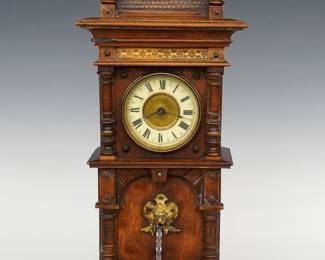 An early 20th century Junghans Fountain novelty clock.  30-hr time only movement with porcelain chapter ring and Brass center medallion, Roman numerals with separate key wind lower movement for the fountain mechanism which runs about one hour, both with Junghans "Crossed Arrow" marks.  Walnut case in the form of a clock tower with three turned finials and arched roof over the clock dial flanked by turned pilasters and a lower "fountain" with revolving twisted glass tube on a shaped bracket base.  Old finish with minor wear, running and functioning when cataloged.  18 3/4" high.  ESTIMATE $600-800