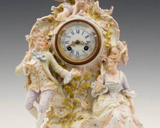A late 19th century Lenzkirch Porcelain Table clock.  8-day spring driven time and strike movement with porcelain dial, and Roman numerals, fancy gilded Brass filigree hands, serial #985120 indicates a production date of 1893.  Louis XV Style figural porcelain case depicts a courting scene with a lady and gentleman beneath a floral arch with Cupid watching from above, scrolled design with applied flowers and hand painted detail with gilded highlights, circular dial door with convex beveled glass.  Slight wear, minor damage, running when cataloged.  16 1/4" high overall. ESTIMATE $800-1,200