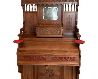 70 - Cornish Company Upright Piano 76"x42"x23" (Description): Cornish Company upright piano featuring  ornate carved wooden cabinetry with  decorative scrollwork and spindle details.  The instrument includes a beveled mirror in  the upper gallery section, two round candle or lamp shelves with red felt covering, and  matching red felt on the foot pedals. The  front panel displays pierced fretwork with  red fabric backing. Measures 76 inches wide  by 42 inches deep by 23 inches tall.