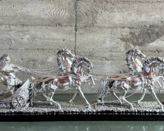 This lot features a Roman Chariot made in Italy and is crafted from silver-coated resin and mounted on a polished black wooden base. This piece measures, 15” H x 44” W x 9” D.