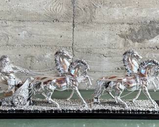 This lot features a Roman Chariot made in Italy and is crafted from silver-coated resin and mounted on a polished black wooden base. This piece measures, 15” H x 44” W x 9” D.