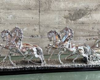 This lot features a Roman Chariot made in Italy and is crafted from silver-coated resin and mounted on a polished black wooden base. This piece measures, 15” H x 44” W x 9” D.