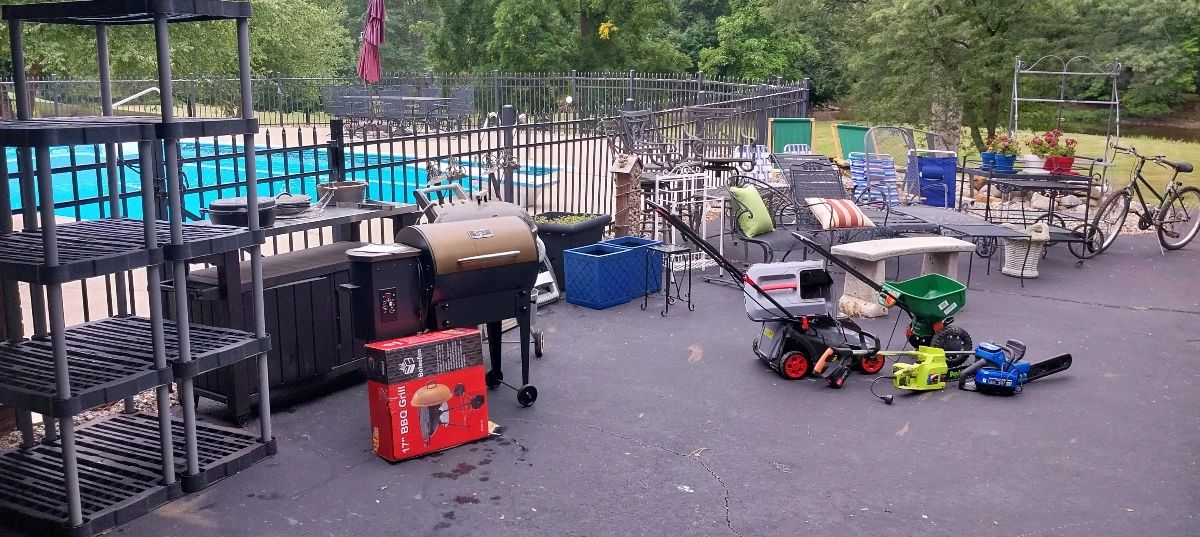 Woodard patio table with 8 spring rocker chairs, offset umbrella, chaise lounges, bar carts, iron bar stools, iron bakers rack, EWork battery operated lawn mower, battery operated string trimmer, Scotts yard spreader, Weber charcoal grill, hose reel, lawn hoses, Himombo wood pellet smoker, stainless top garden cart, metal shelving, plastic shelving, Kobalt 40v chainsaw, Poulan electric chainsaw, cement bench, Diamondback mountain bike, Tommy Bahama beach chairs, cement basket