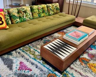 Green tufted mid-century sofa with floral velvet throw pillows, matching green ottoman, low wood coffee table with built-in backgammon board, colorful shag area rug, coffee table books.