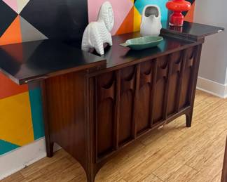 Walnut Brutalist-style credenza with sculpted geometric door panels and pull-out leaf, red glazed ceramic vase, white ceramic elephant figurine, sage green ceramic dish, white geometric pitcher.